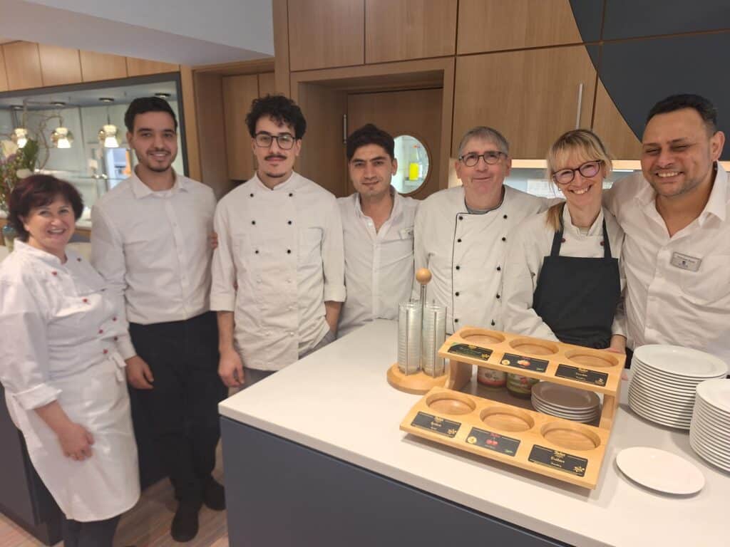 Gruppe von Mitarbeitern in Kochkleidung, die an einem Tisch in der Küche stehen. 01 M.