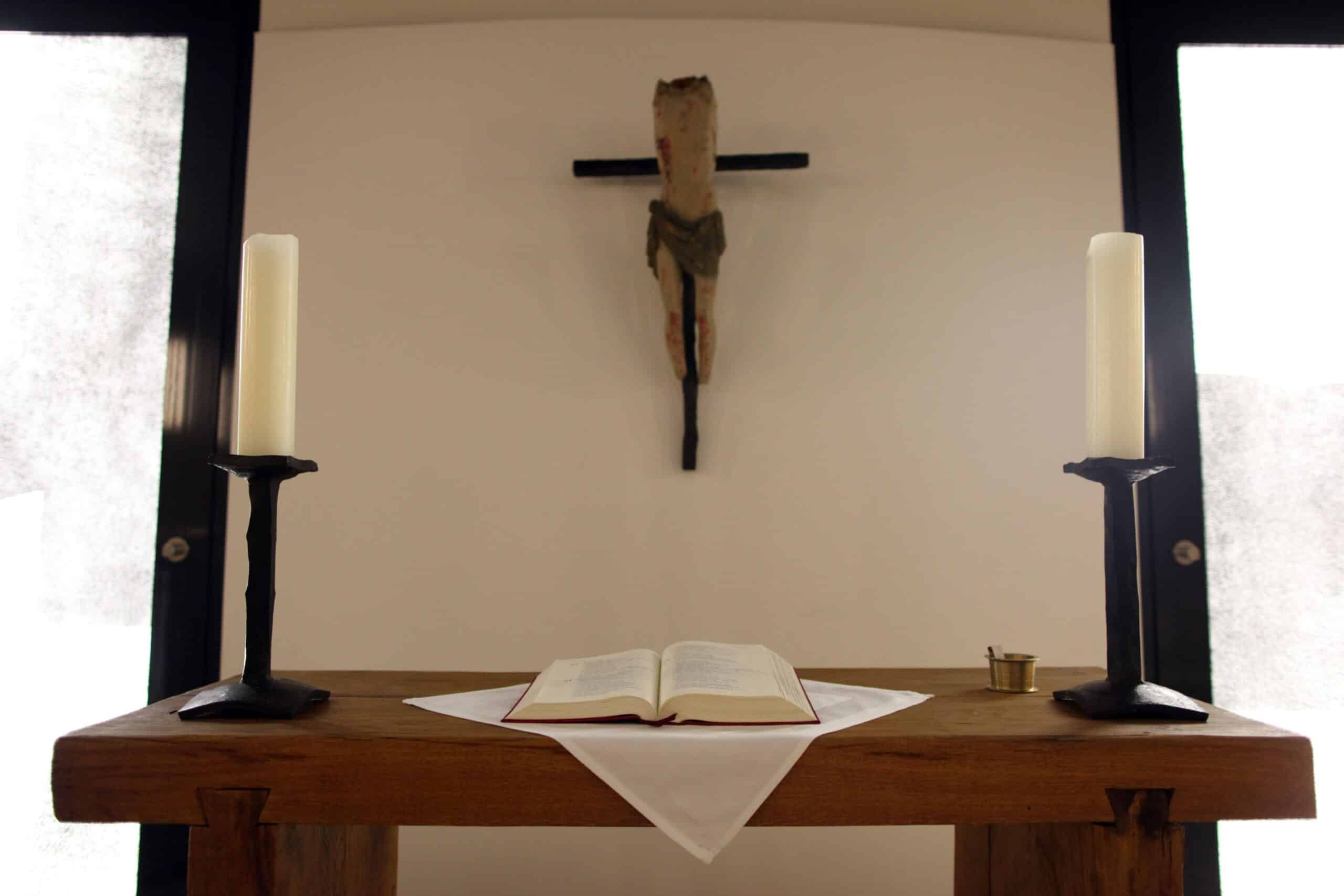Ein Holztisch mit einer offenen Bibel, zwei Kerzenhaltern und einem Kreuz im Hintergrund.