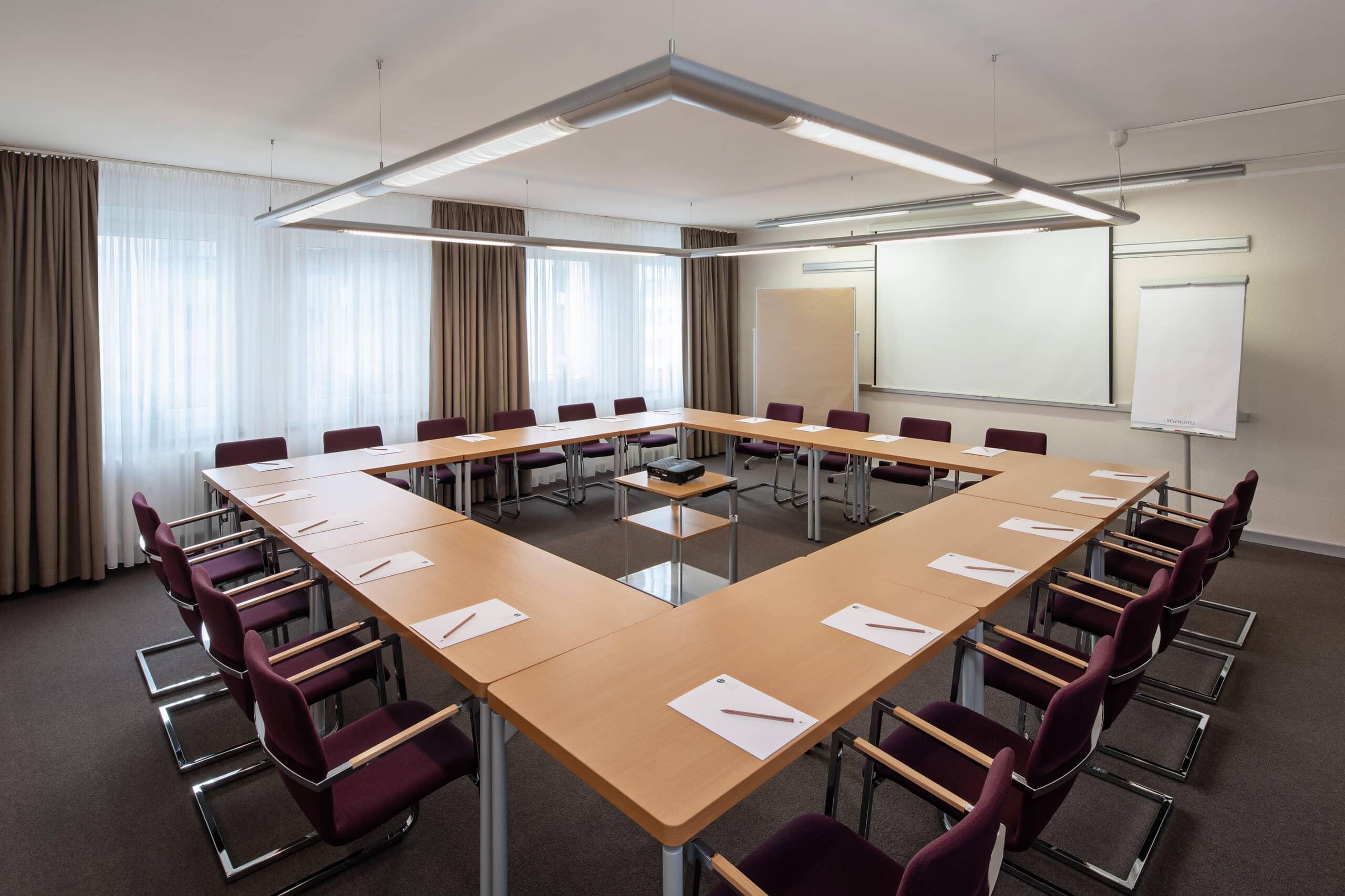 Konferenzraum mit einem großen Tisch in U-Form, umgeben von Stühlen und ausgestattet mit Präsentationstechnik.