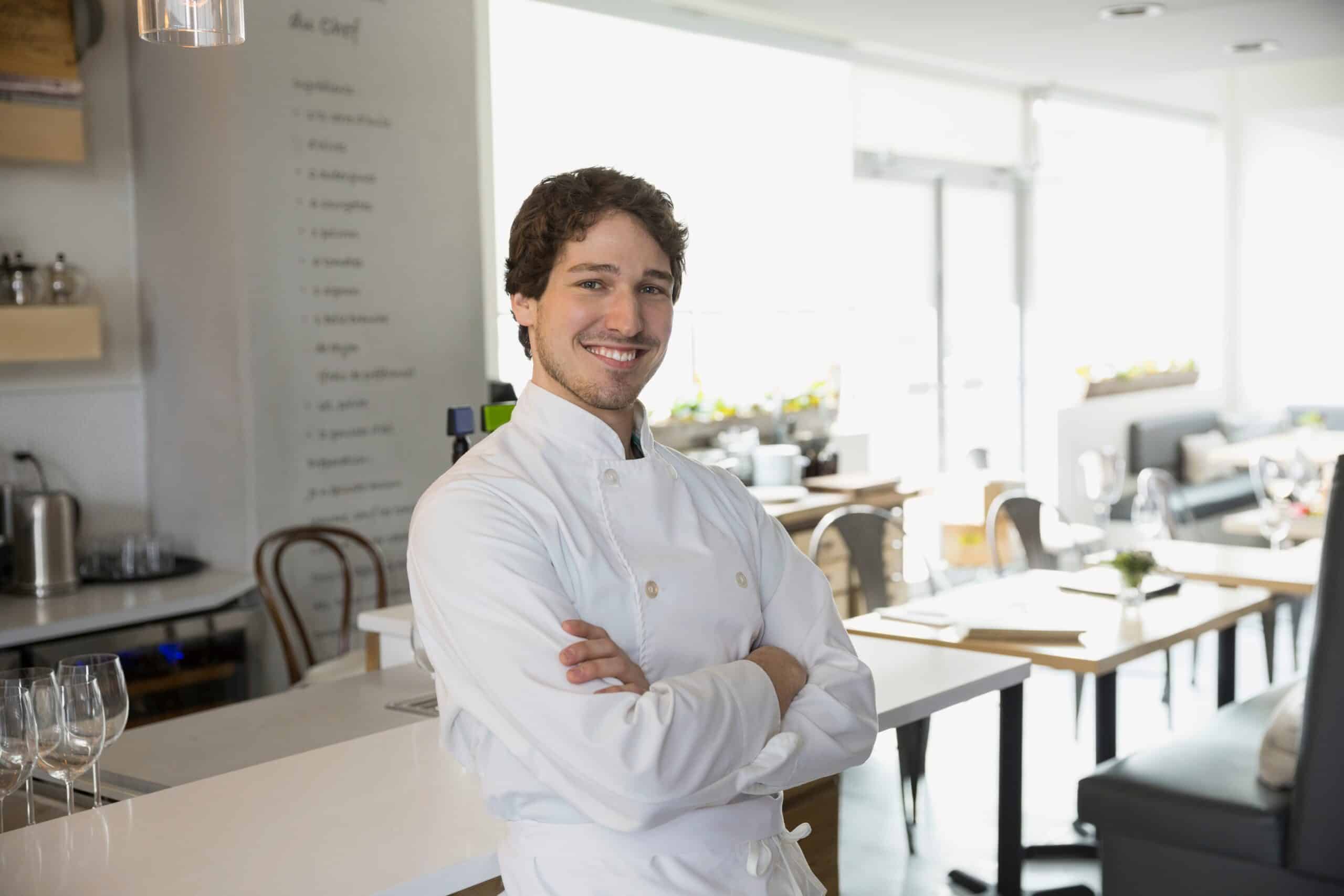 Ein selbstbewusster Bistro-Chef in weißer Kochjacke mit verschränkten Armen lächelt in einem modernen Restaurant.