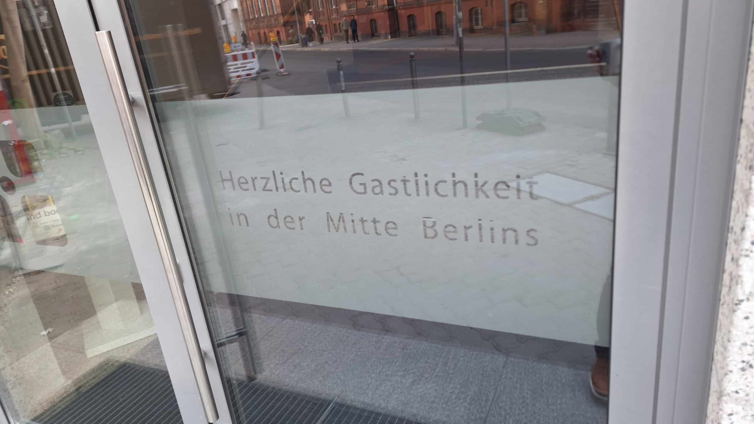 Transparente Tür mit der Aufschrift 'Herzliche Gastlichkeit in der Mitte Berlins' in einem modernen Gebäude.
