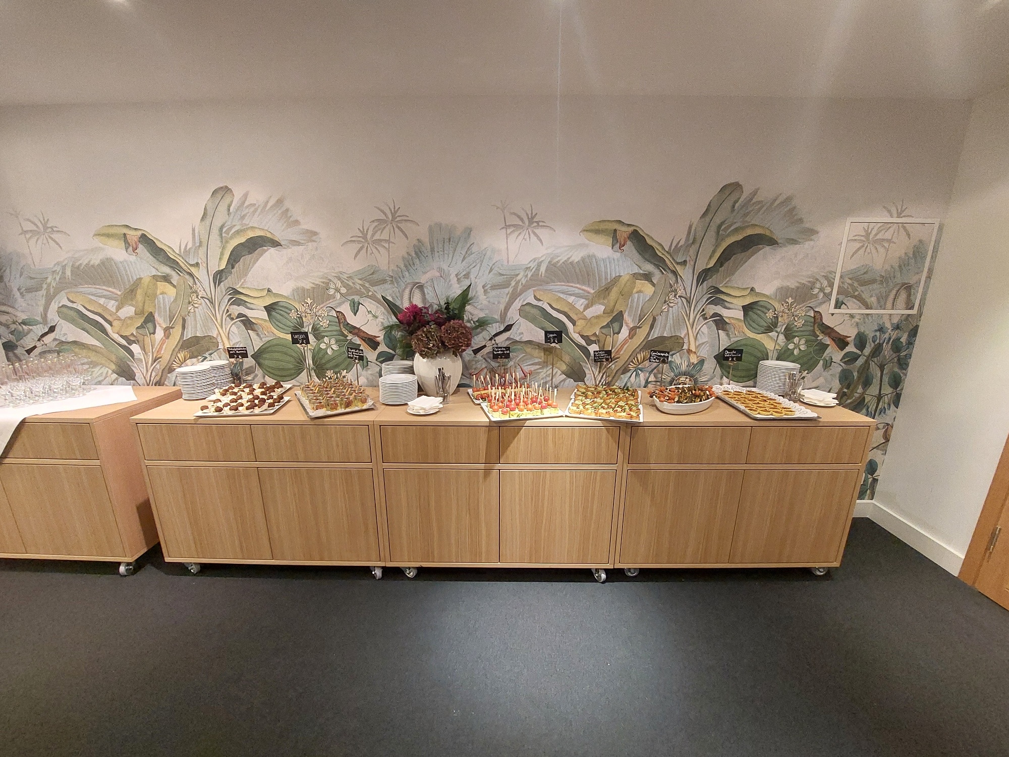 Buffet mit verschiedenen Speisen auf einem Holztablett in einem modernen Raum mit Pflanzenmuster an der Wand.