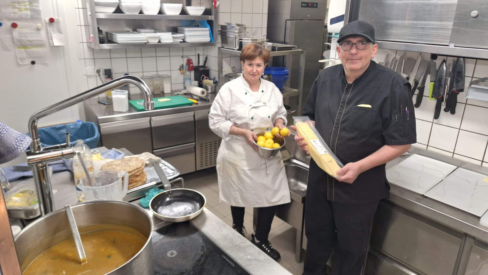 Zwei Köche in einer Hotelküche, einer hält frische Zitronen, der andere eine Packung Pasta.