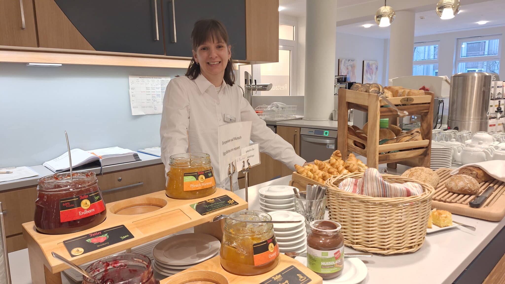 Eine Mitarbeiterin präsentiert eine Auswahl an Marmeladen und Brötchen in einem Buffetbereich.