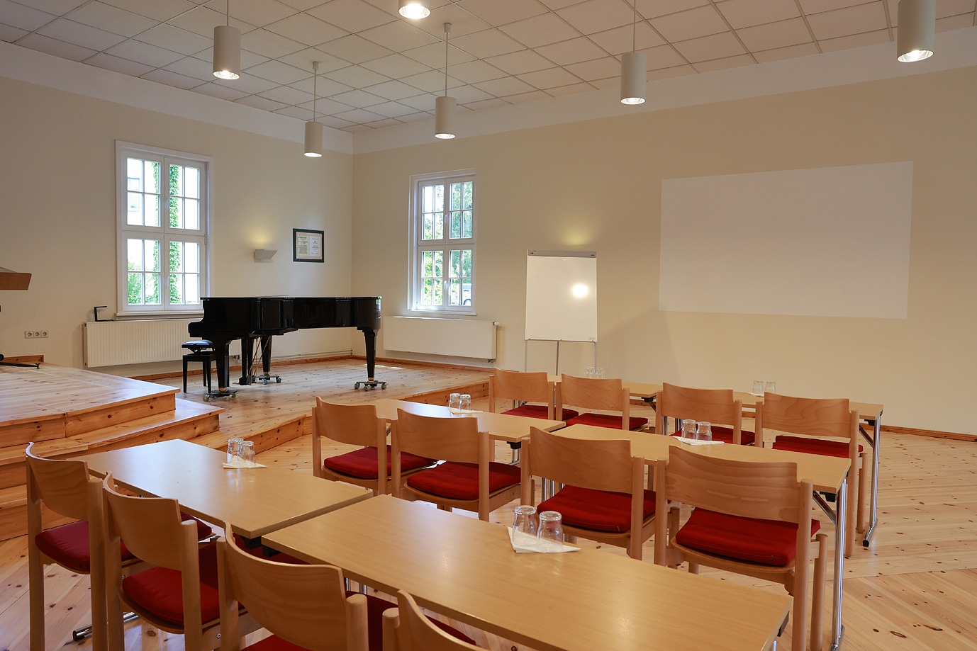 Ein schwarzer Flügel steht in einem Raum mit Tischen und Stühlen, bereit für eine Veranstaltung im Hotel Dietrich Bonhoeffer Haus.