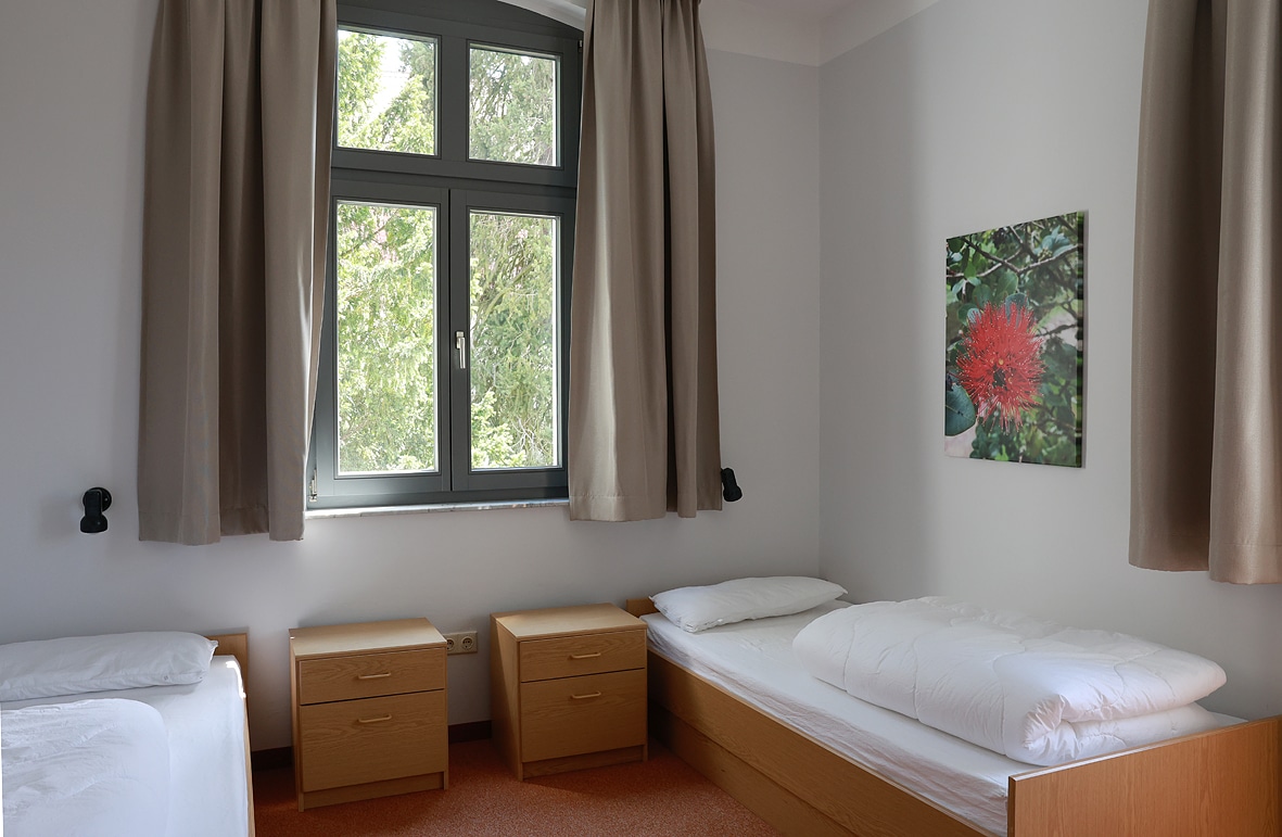 Zimmer mit zwei Einzelbetten, Holzkommoden und einem Fenster mit Vorhängen.